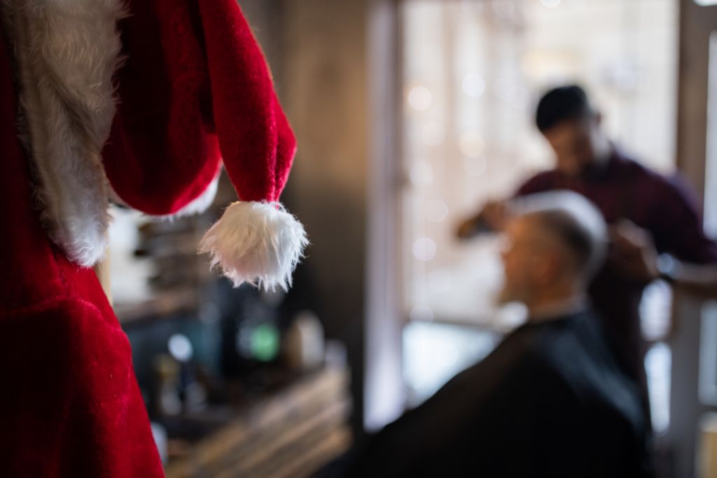 In primo piano: cappello di babbo natale appeso.
Sfondo: barbiere che tagli i capelli al cliente